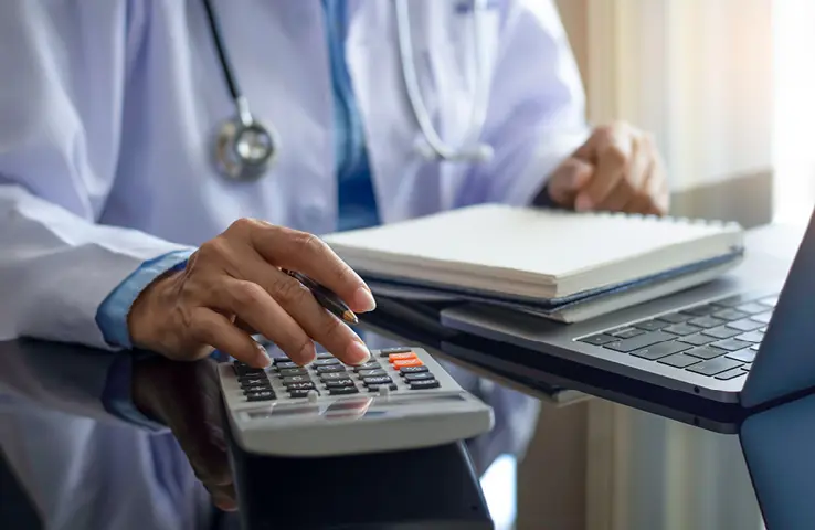 Le médecin est assis à la table devant un bloc et un ordinateur portable et tape sur la calculatrice.