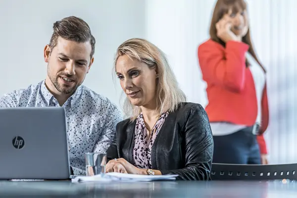 Zwei Aussendienstmitarbeitende sitzen am Tisch und arbeiten am Laptop; Eine weitere Aussendienstmitarbeiterin steht im Hintergrund und telefoniert